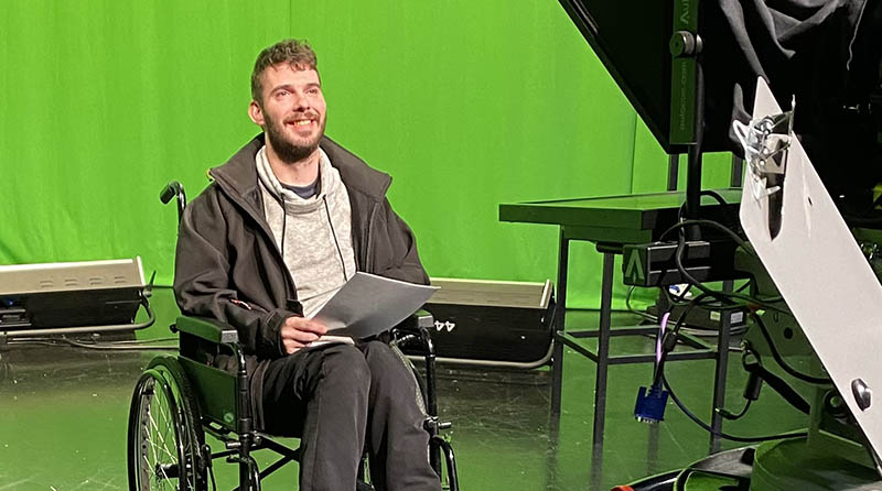 A student in a wheelchair with green screen behind them, filming a segment in the university TV studio