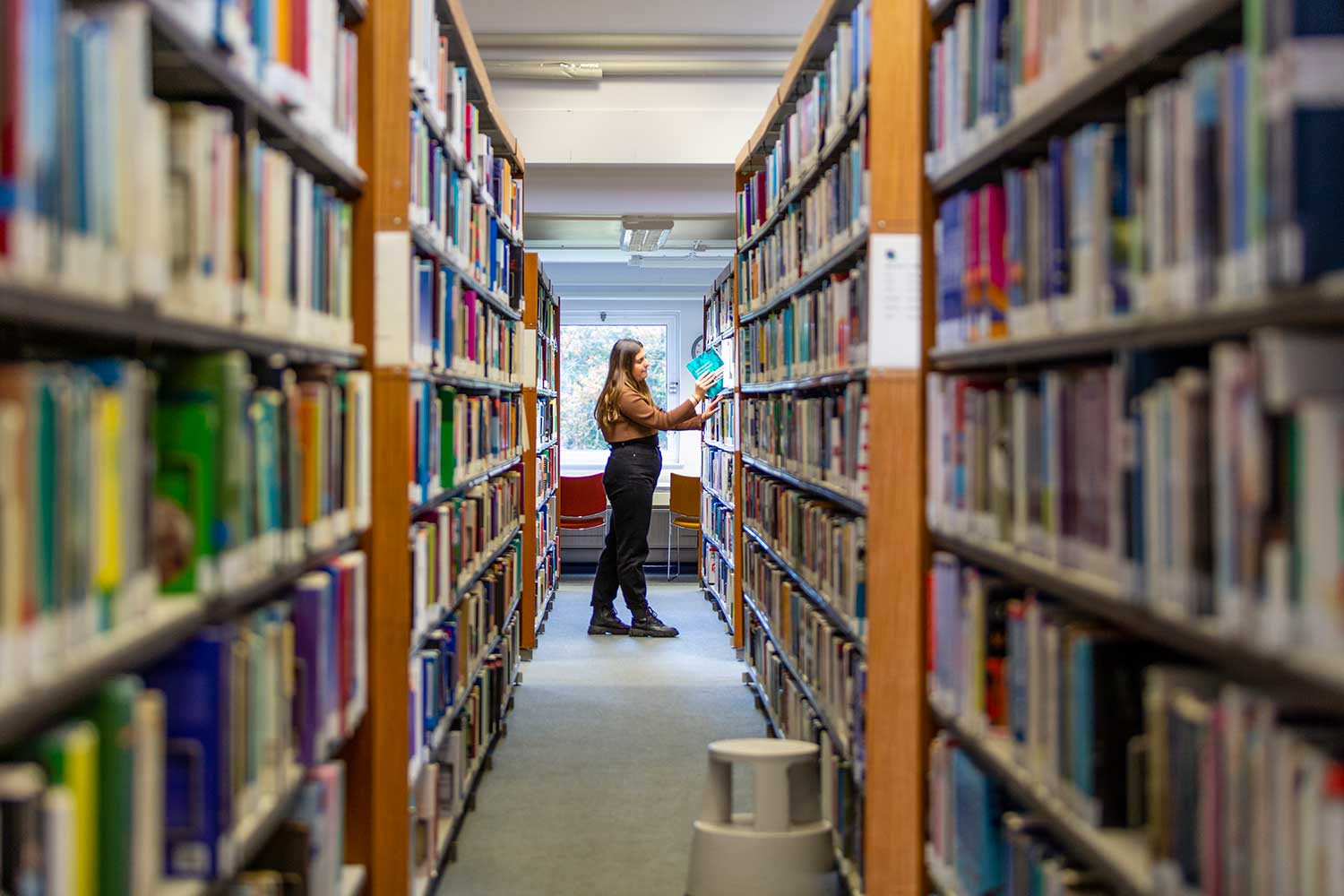 A woman stands between two high rows of library shelves browsing the books on the shelf in front of her