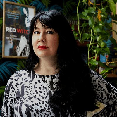 Anna McKerrow's profile photo. She is stood in front of a framed cover of one of her novels, Red Witch.