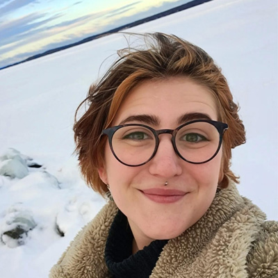 Person with short hair wearing glasses smiling at camera against background of snow-covered countryside