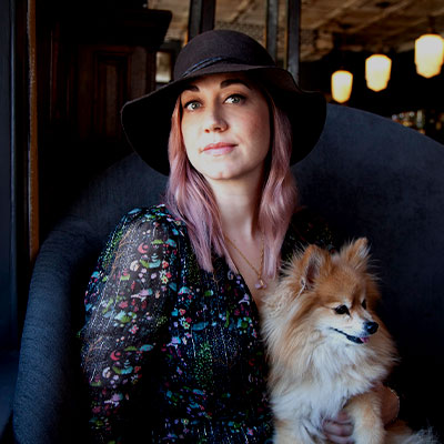 Confident woman with pink hair wears a hat holds a small dog on her lap