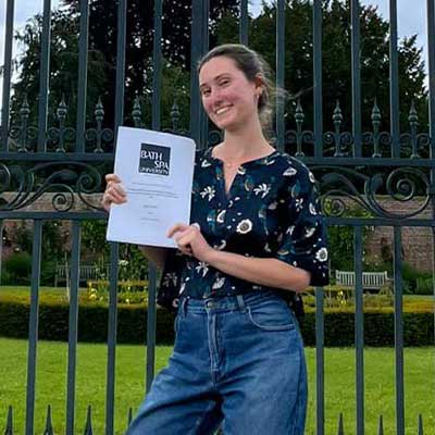 A smiling student stands at the gates of the Italian Gardens at Newton Park and holds her dissertation