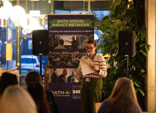 person talks into a microphone in front of a pull up banner saying “Bath Social Impact Network”, addressing a crowd facing them.