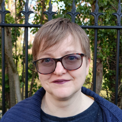 Person with short fair hair and wearing blue jumper smiling in front of a park railing