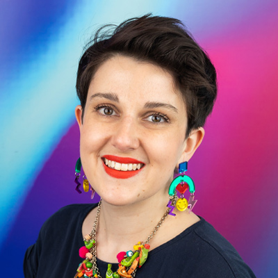 A smiling Bath Spa University staff member sits in front of a brightly washed background of blue and purple.