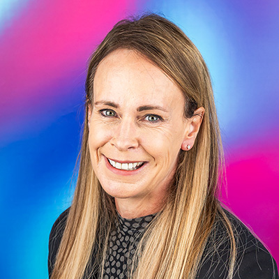 A photograph of a smiling staff member with long hair against a blue and pink background.