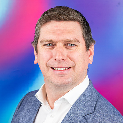 A smiling Bath Spa University staff member sits in front of a blue and pink background