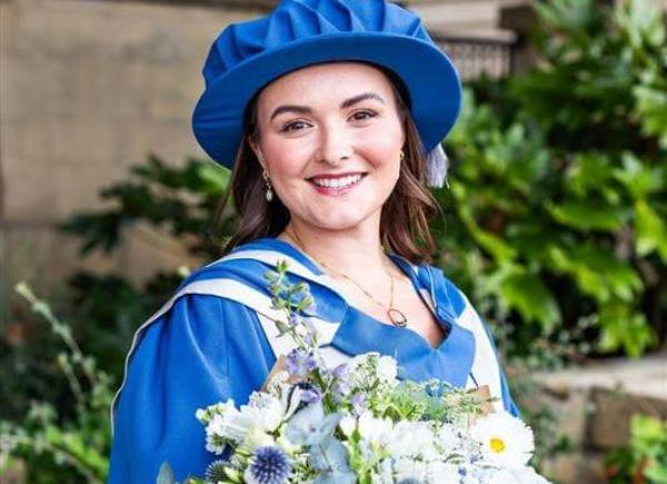 Lou May wearing Honorary Doctorate clothing, holding large bouquet of flowers