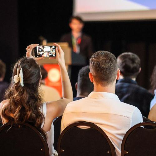 A person stands on stage with a large screen behind them and an audience of people looking their way. A member of the audience has their phone up in the air taking a photo.
