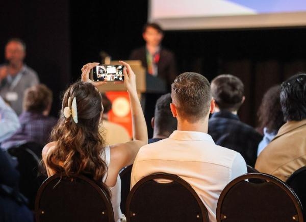 A person stands on stage with a large screen behind them and an audience of people looking their way. A member of the audience has their phone up in the air taking a photo. 