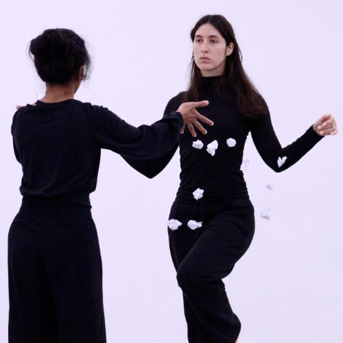 Two female dancers facing each other, one with leg lifted. Both wearing black.