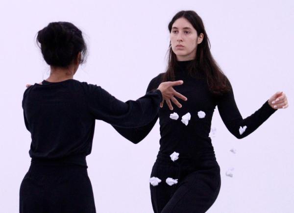 Two female dancers facing each other, one with leg lifted. Both wearing black.