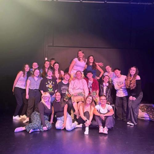 A group of musical theatre society members clump together for group photo, under a spotlight, against a black wall