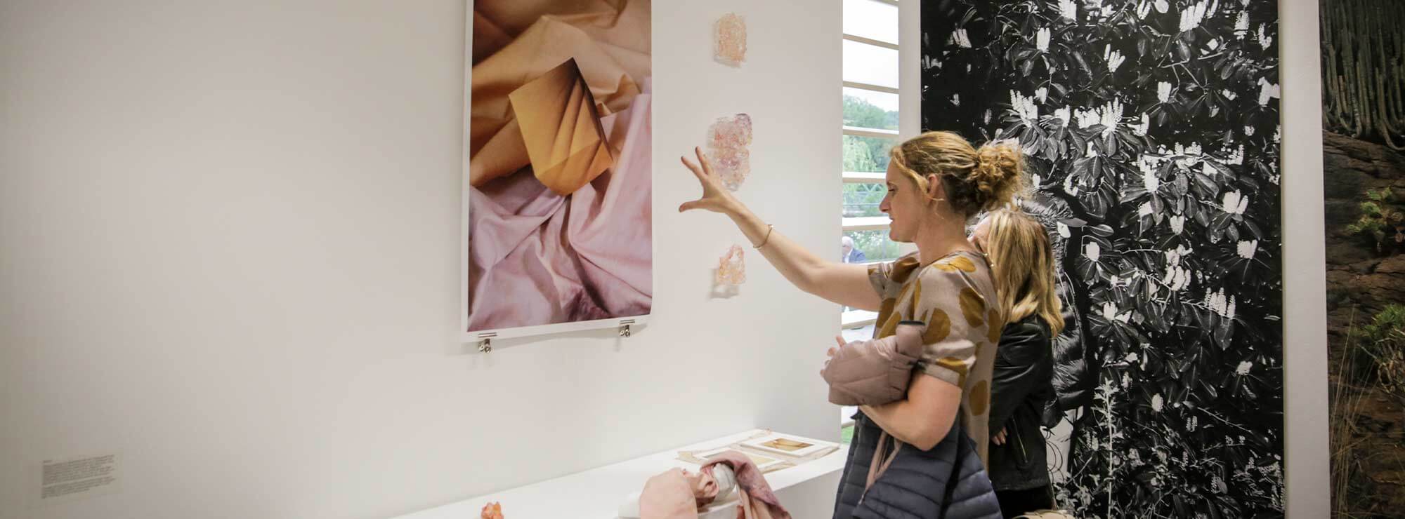 Person with long blonde hair standing in front of a painting at a degree art show