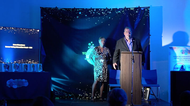 A man on the right wearing a grey suit and a purple shirt stands behind a lecturn, presenting the awards and to his left a woman wearing a black and white dress is co-presenting