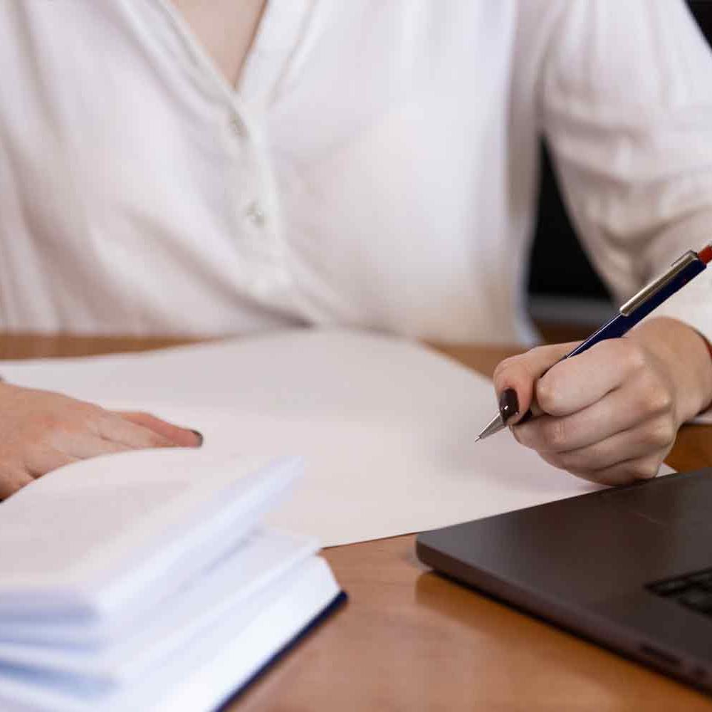 Person in white blouse taking notes with pen and paper from a laptop