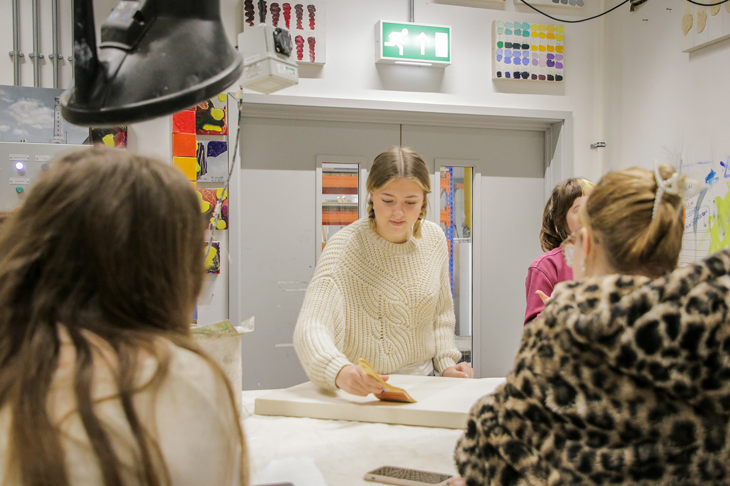 In a workshop in a creative space, a student uses a brush to paint on a canvas on a table while other students watch