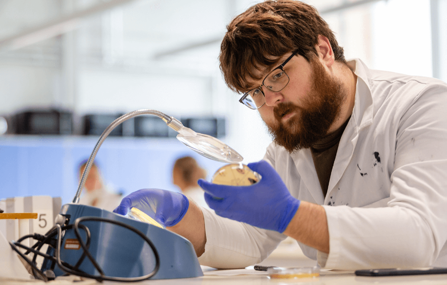 a student in a lab coat, goggles and gloves looks at a petri dish and writes observations