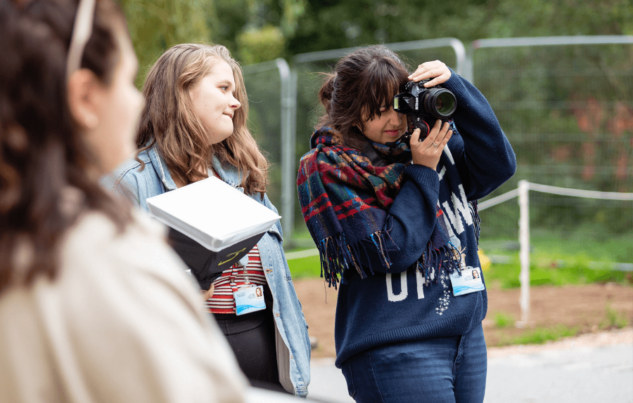 three students stand outside. One is holding a flash box and another is taking photos.