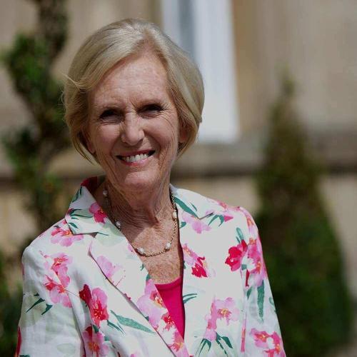 Woman smiles to the camera wearing a floral jacket. Formal topiary and building are in the background