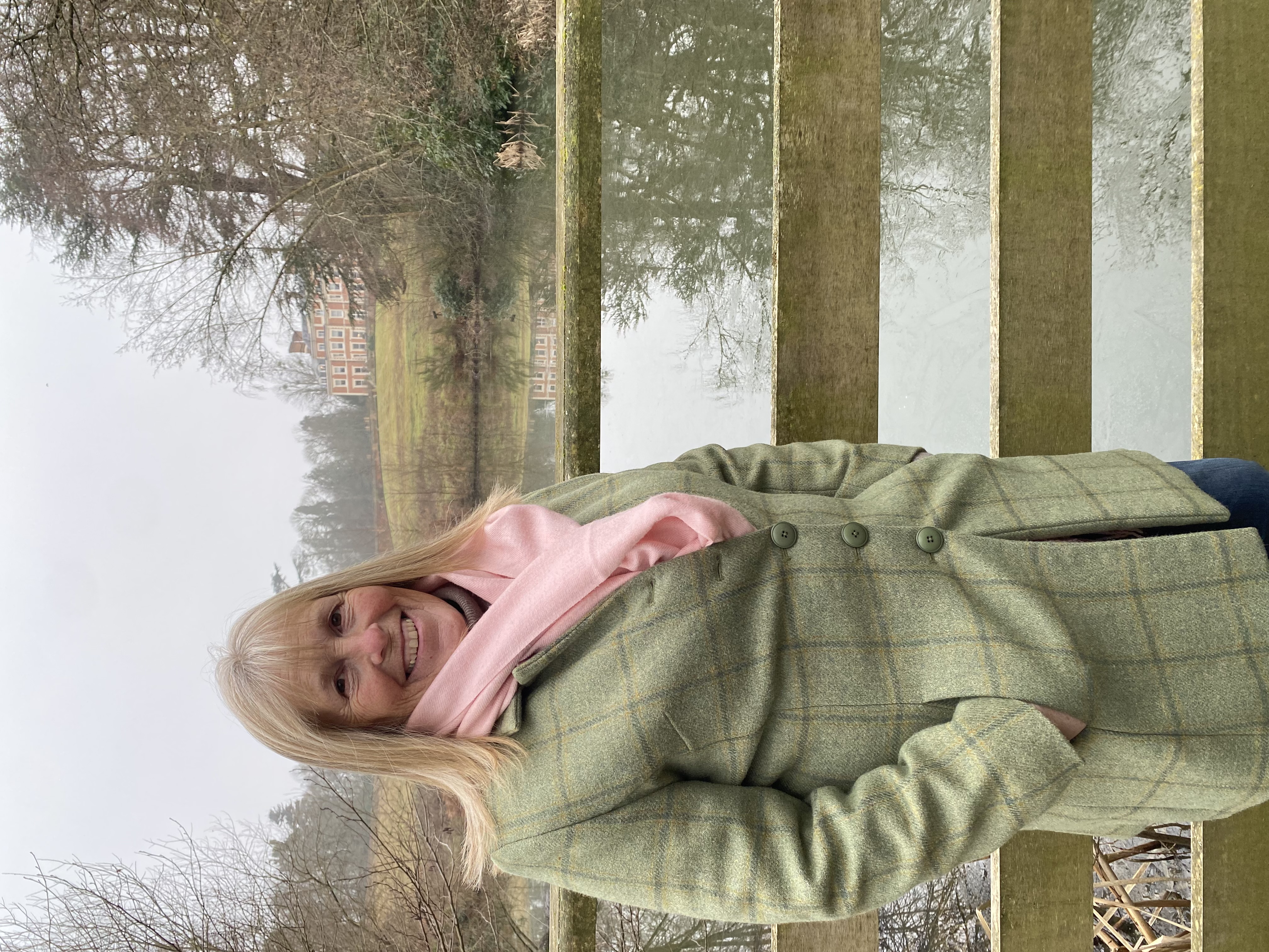 Blonde woman in winter jacket and scarf stood by scenic lake