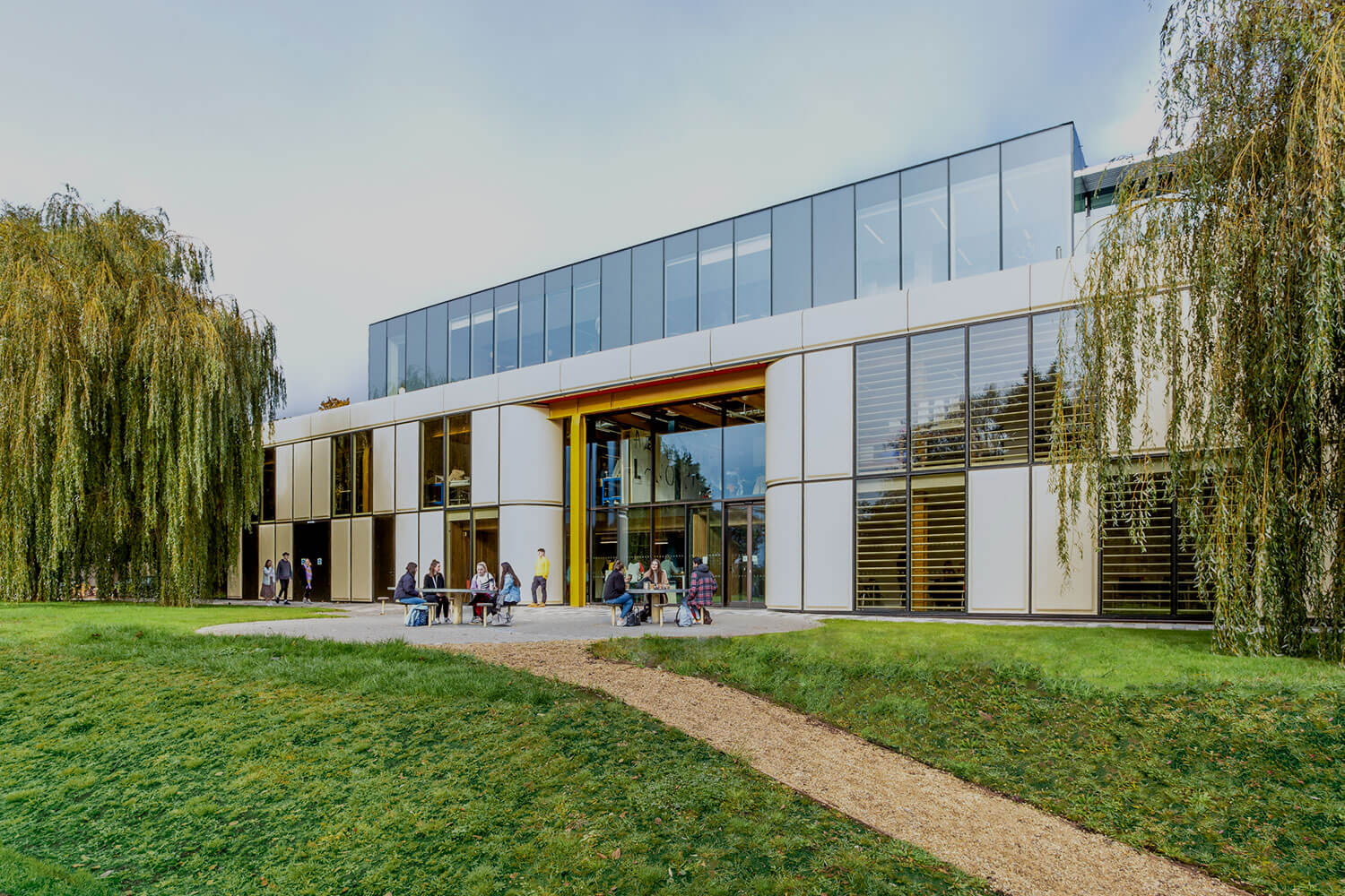 Exterior of a period factory building with modern glazing design, flanked by mature willows, with students in the outdoor seating area