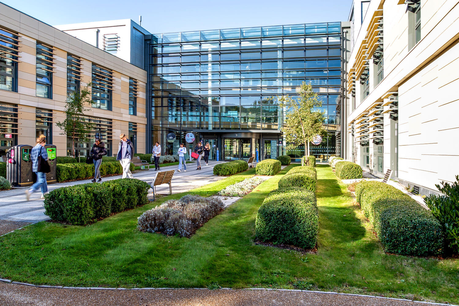 A modern, glass-fronted building with a garden out front and students walking past