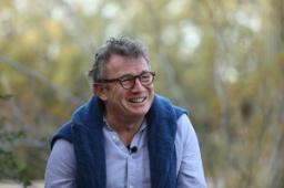 A man wearing glasses and a blue jumper over his shoulders with outdoor background