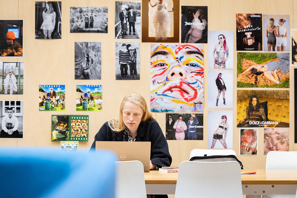 A student sat at a laptop in Locksbrook Campus, a wall behind shows lots of student artwork