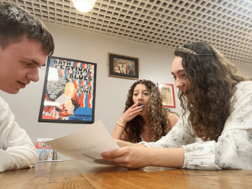 Three students in regency dress sit around a table looking shocked at a letter.
