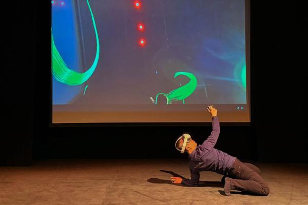 A dancer performs on stage wearing a VR headset and motion tracked controllers. Traces of colour on the screen behind the dancer show the path of their motions.