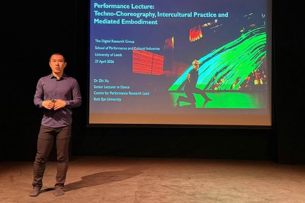A professor stands on a stage standing in front of a slide showing the title of the lecture: 