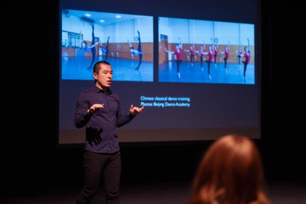 A lecturer on stage stands in front of a slide labelled 