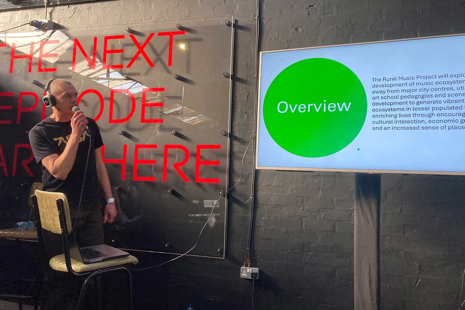 A person stands in front of a dark wall and neon signs to present to a group