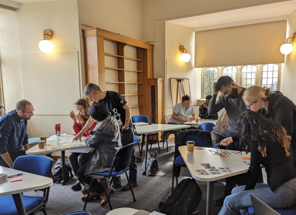 A room of students stood around three tables, working on tasks about data analytics.