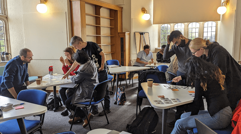 A room of students stood around three tables, working on tasks about data analytics.