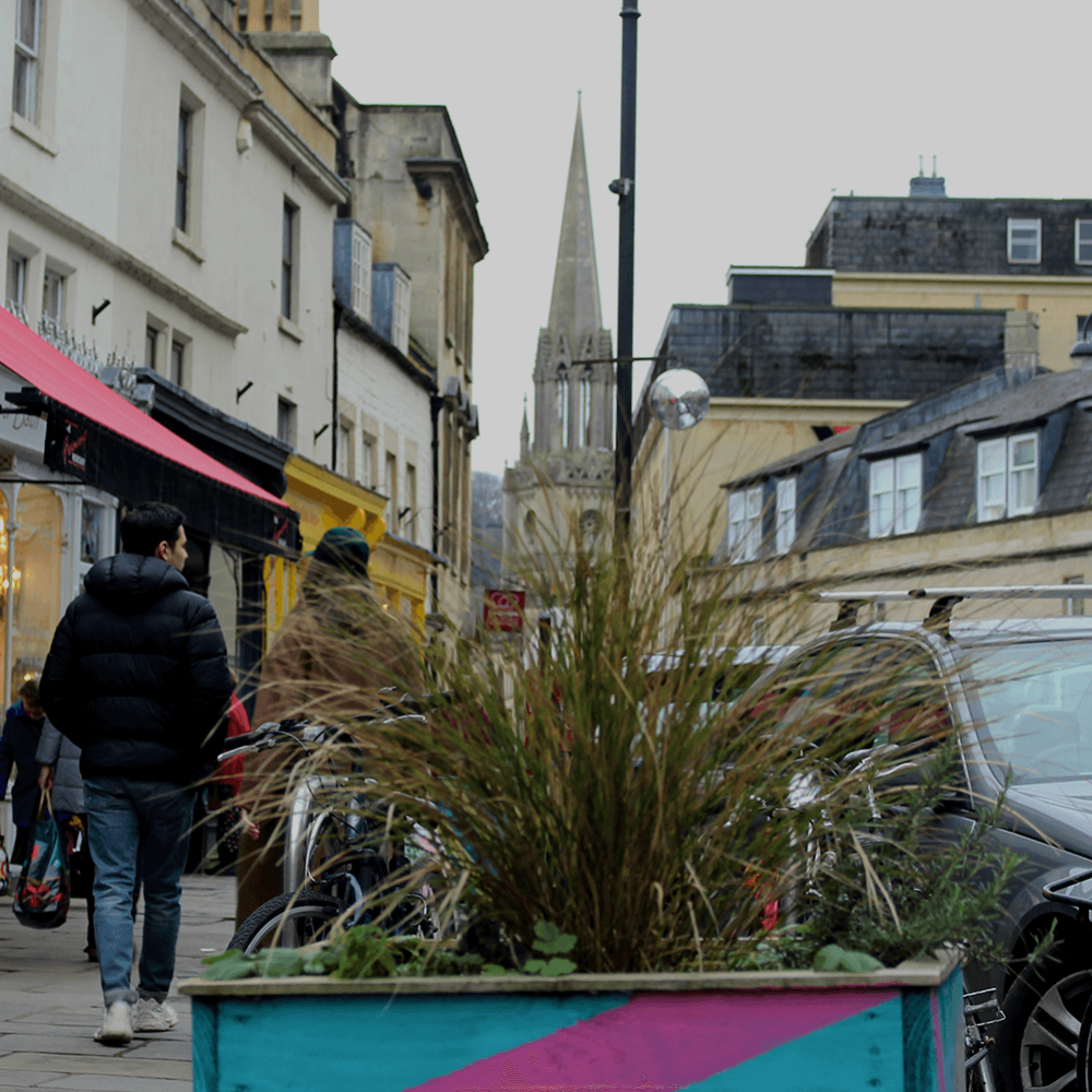 In the foreground of the image is a plant in a blue and purple striped planter. Behind this is a row of shops on walcott street. In the distance is the top of a church spire