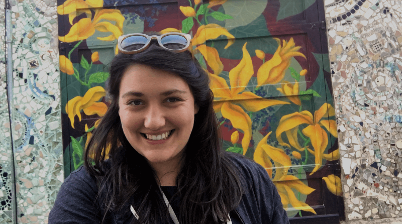 Lior Burstein standing in front of a mosaic wall. She stands in front of two doors that are painted to look like flowers.