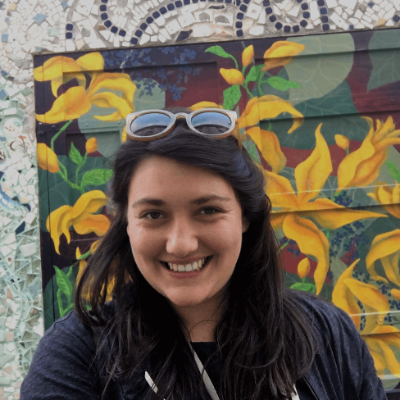 Lior Burstein standing in front of a mosaic wall. She stands in front of two doors that are painted to look like flowers.