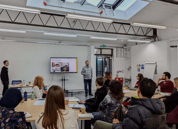 a group of students sit round tables listening. at the back of the room a person stands giving a lecture.