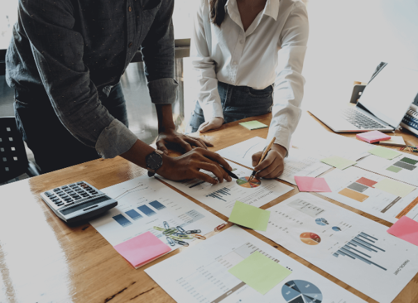 Two coworkers stand next to a table. On the table are sheets of paper showing different diagrams of data. They are both leaning over the sheets to point at one graph.