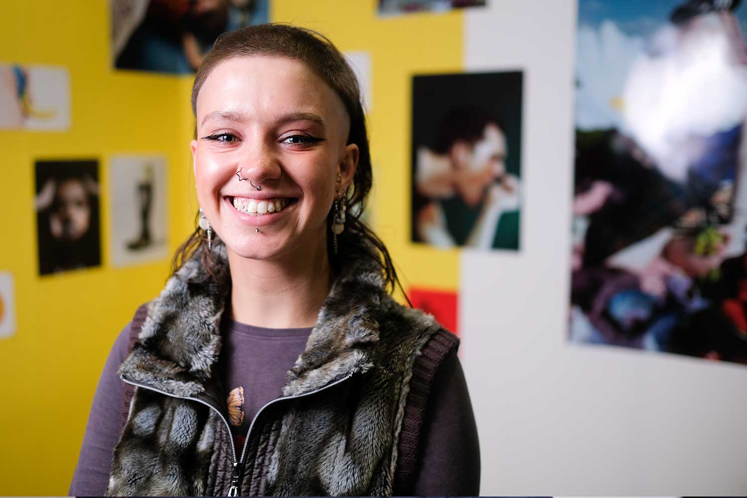A person with bold eyeliner and several piercings smiles at the camera