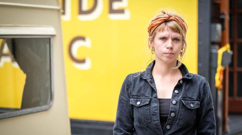 Woman wearing overalls and a hair scarf stands next to a painted caravan