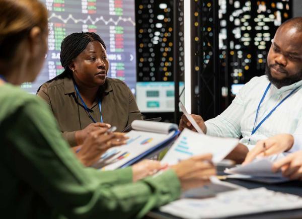 Three people seated at a meeting room table with documents on the table and on graphs displayed on digital screens