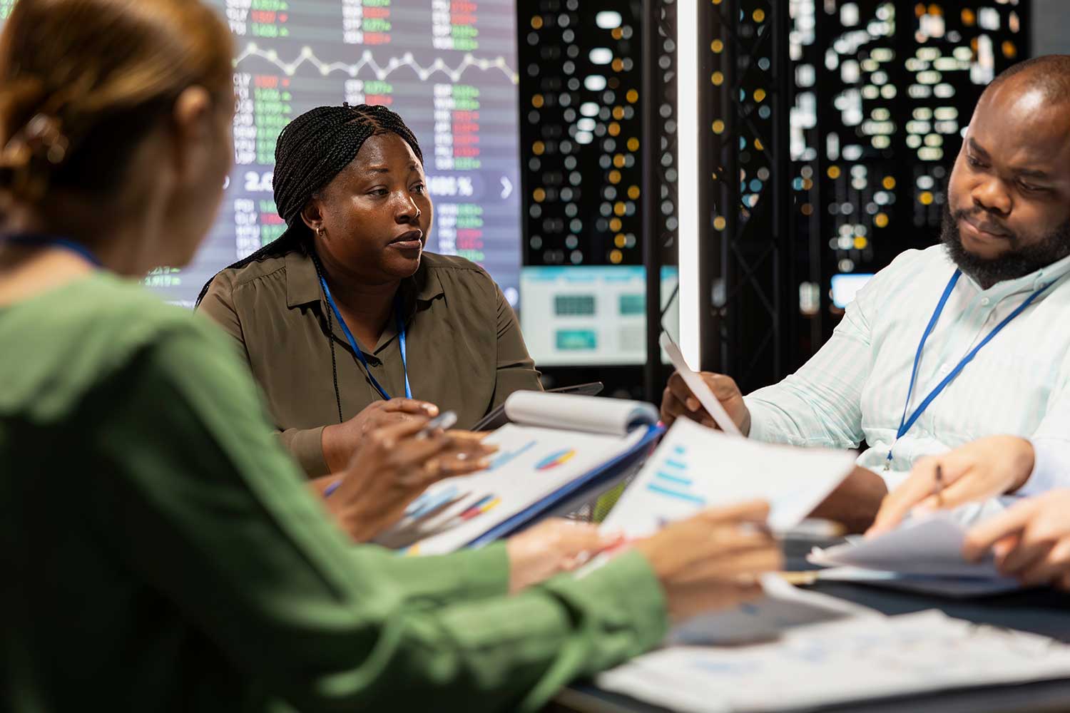 Three people seated at a meeting room table with documents on the table and on graphs displayed on digital screens