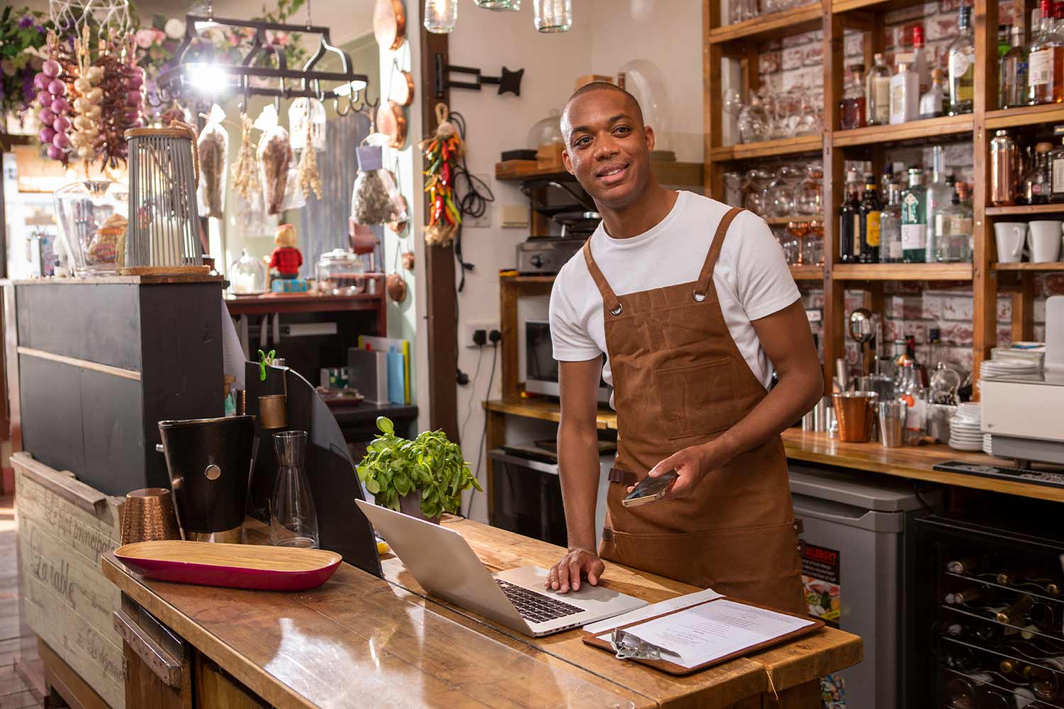 POC standing in a cafe wearing an apron looking over laptop and pages of accounts