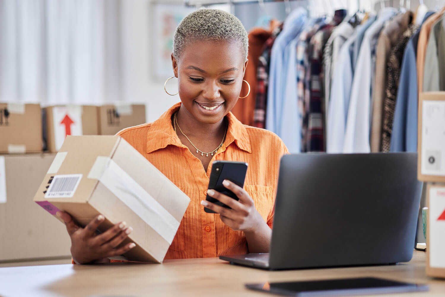 Woman with laptop, phone and parcel and a clothing rail in the background
