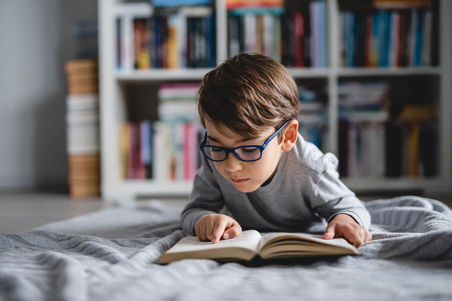 A child wearing glasses lying on a bed and reading a book, using his finger to track the words