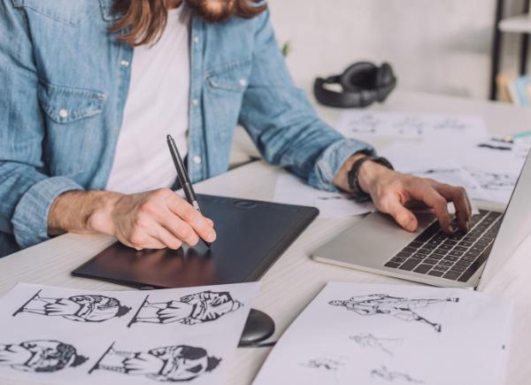 Person in denim shirt in a workshop drawing animation by hand while using computer