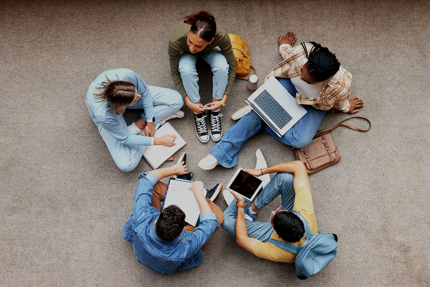 A group of students sitting on the floor with laptops, notebooks and tablets.